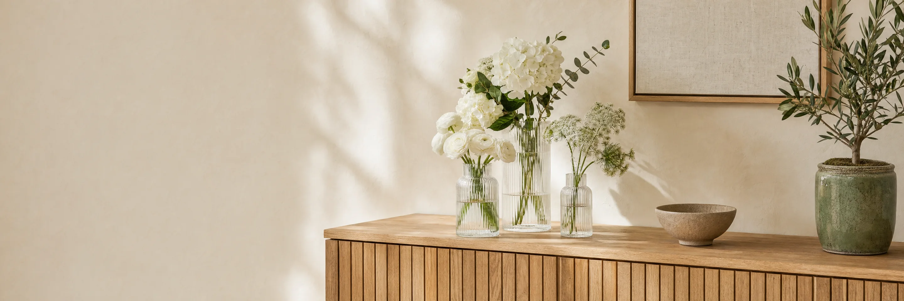 Drei Rillenglasvasen mit weißen Blumen, eine Schale und ein Olivenbäumchen auf einem Eichen-Sideboard